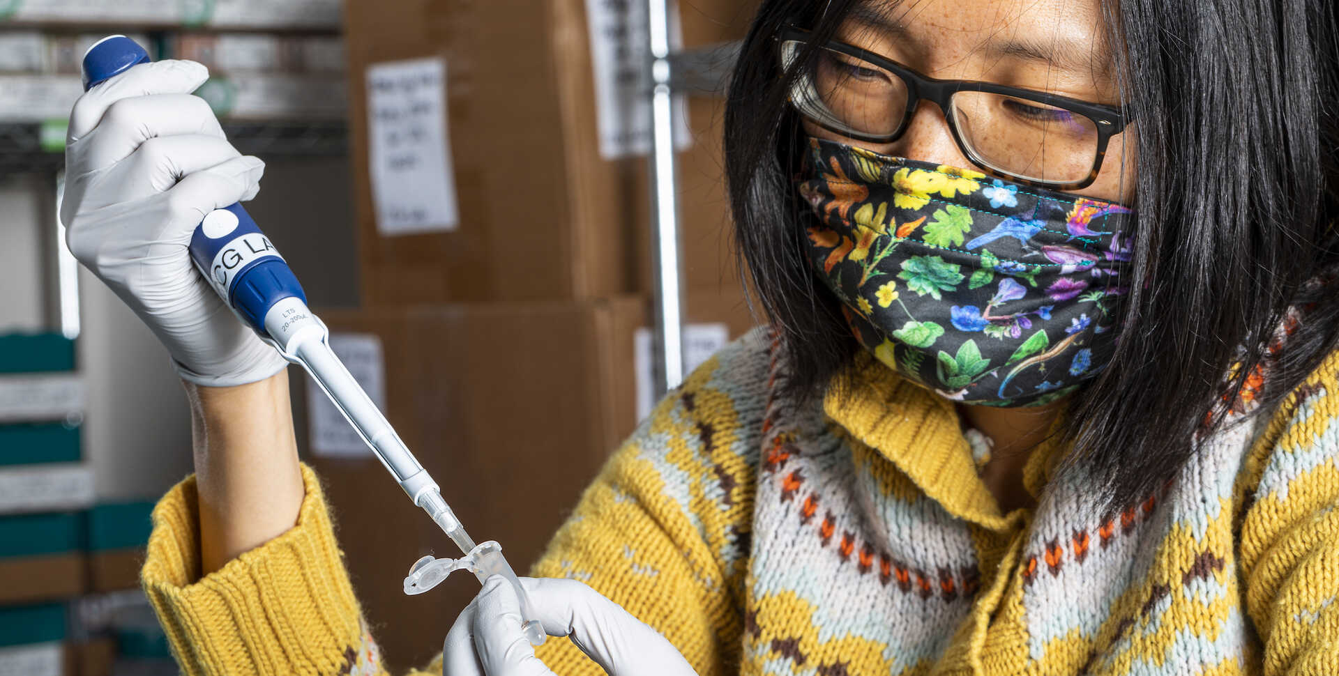 Athena Lam, wearing a rainbow mask and gloves, extracts DNA using a blue pipette tool. 