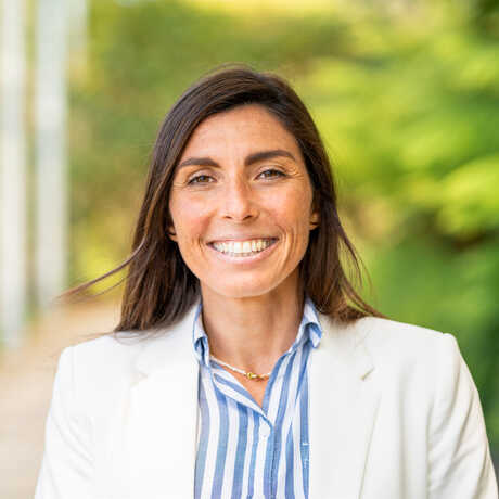 Estefanía Pihen González, Chief of Education and Learning is pictured wearing a blue and white striped shirt and white blazer in front of green plants. 