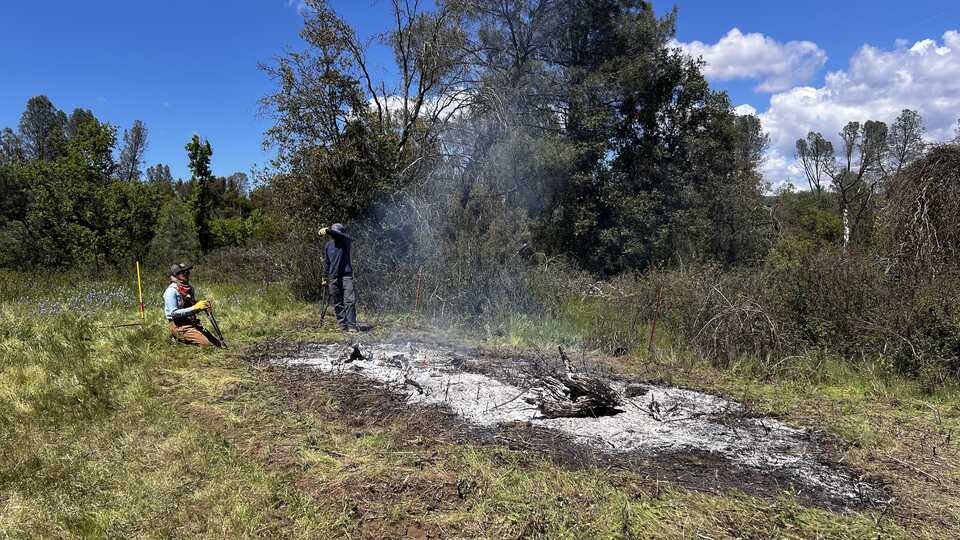 Two people preside over a smoldering pile of white ashes, evidence of a recently completed burn. 