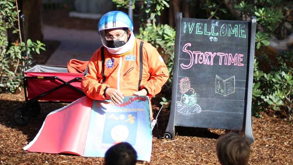An educator dressed as an astronaut reading a storybook outside