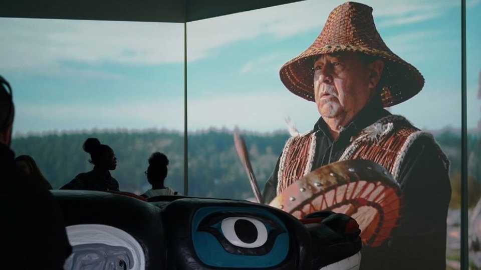 House of Tears Carver of the Lummi Nation pictured behind a carved totem, with guests observing